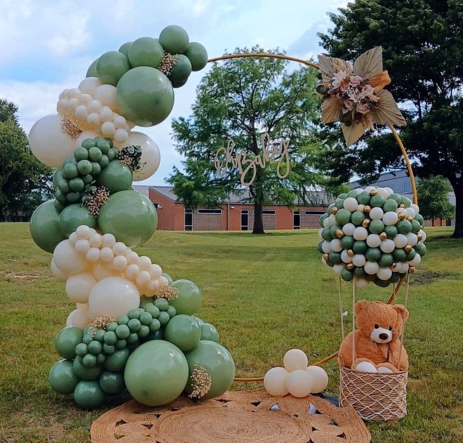 Sage green and cream balloon hoop arch with a teddy bear basket set up outdoors for a baby shower