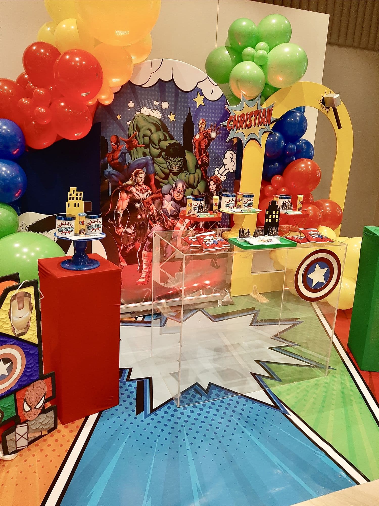 Wide view of a superhero themed birthday setup with balloon garlands, comic backdrop and dessert table