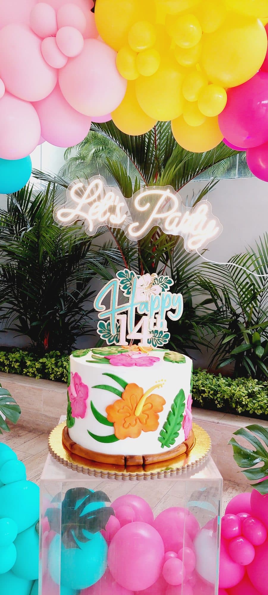 Close-up of a tropical fruit-themed birthday cake in front of a pink, teal and yellow balloon wreath