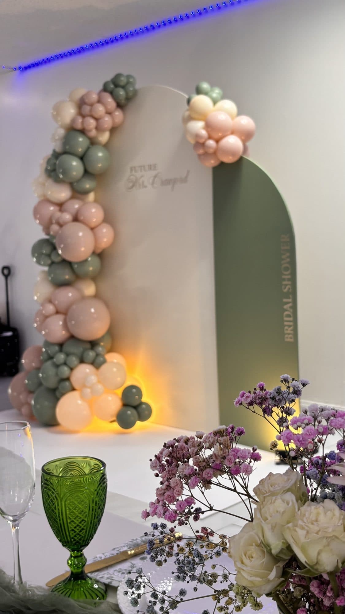 Detail of a sage green, blush and cream balloon arch beside a green glass vase with flowers