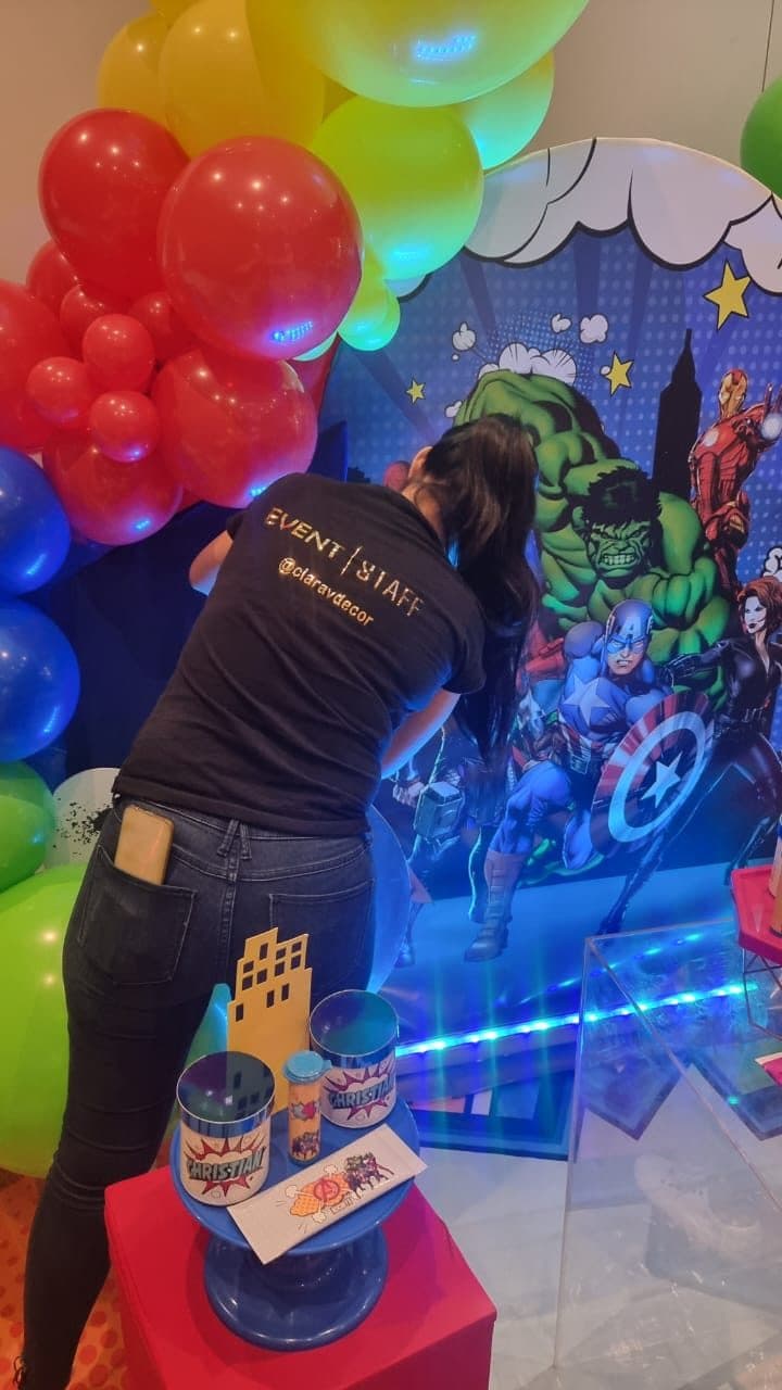 Clara Vento adjusting balloons on an Avengers themed birthday backdrop, wearing a ClaravDecor event staff shirt