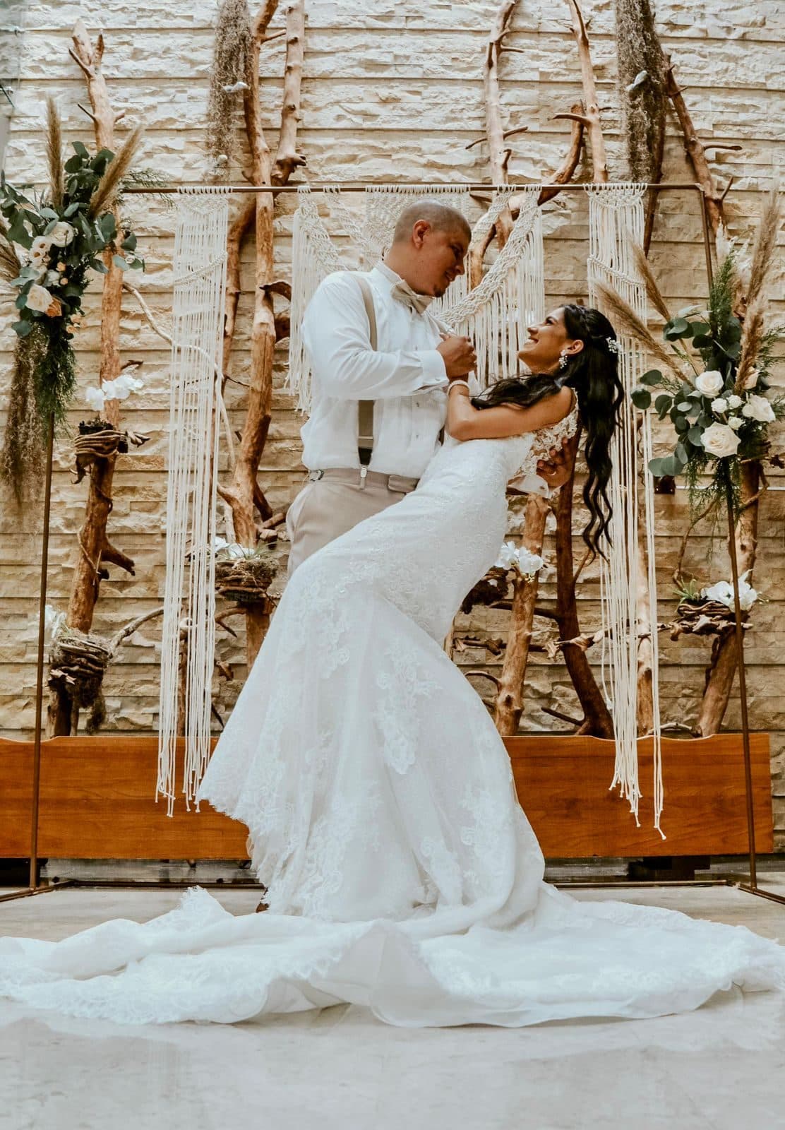 Bride and groom embracing under a boho macrame ceremony arch with driftwood branches, pampas grass and white florals